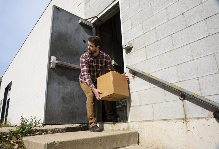 Loos Prevention topic. A male warehouse employee placing a box outside a back door of an industrial warehouse distribution building. A typical internal employee theft scenario.